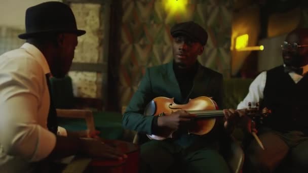 アフリカ系アメリカ人の男性ジャズトリオのミディアムショットは 楽器と円に座って 居心地の良い暖かい薄暗いライトでクラブでの夜のコンサートをリハーサルしながら — ストック動画