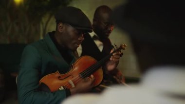 Koyu yeşil takım elbiseli, bereli, bando provası sırasında keman çalan yetenekli Afrikalı genç Amerikalı caz müzisyeninin belden yukarısını çek.