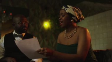 Siyahi kadın caz şarkıcısının şarkı sözlerine bakıp şarkı söylerken, siyahi erkek davulcunun sopalarla vuruşunu, loş ışıklı gösterinin provasını yaptığı orta boy bir sahne.