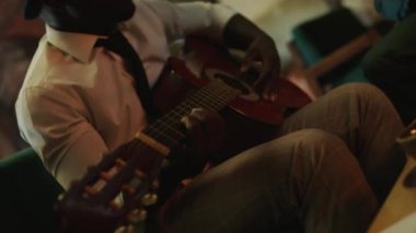 Beyaz gömlekli, siyah kravatlı ve şapkalı genç Afrikalı Amerikalı erkek caz gitaristi akustik gitar çalarken süslü caz kulübünde akşam gösterisine hazırlanıyor.