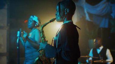 Kadın saksafoncu, vokalist ve erkek bateristten oluşan Afrikalı Amerikalı caz grubunun orta boy çekimi neon sahnede canlı müzik konseri veriyor.