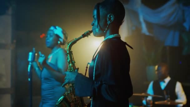 女性のサックス奏者 ボーカリスト 男性ドラマーで構成されたアフリカ系アメリカ人のジャズバンドのミディアムショット クラブのネオンステージでライブミュージックコンサートを行います — ストック動画