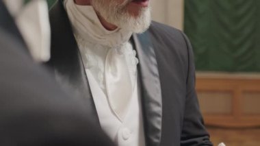 Yakışıklı, olgun, gri sakallı, siyah silindir şapkalı, elleri beyaz eldivenli, Victoria tarzı top partisi görevlileriyle konuşan adam.