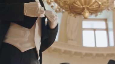 Regency tarzı balo salonunda altın sarısı bir avizeyle dikilirken smokin, yelek ve eldivenleri ayarlayan tanınmamış erkek sosyetenin orta kısmı.