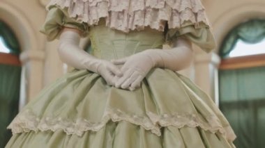 Düşük açılı genç kızıl saçlı beyaz kadın portresi. Büyük yeşil fırfırlı Regency elbisesi giymiş. Altın avizenin altında elleri katlanmış kameraya bakıyor.