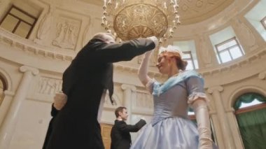 Düşük açılı iki beyaz çift Viktorya dönemi elbiseleri giyip lüks altın avizenin altında dans ederken geniş balo salonunun yüksek tavanında.