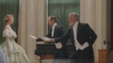 Zarif smokinli ve eldivenli iki yakışıklı beyaz adamın orta yavaş çekimi. Sosyete kızlarına balo partisinde onlarla dans etmelerini teklif ederken.