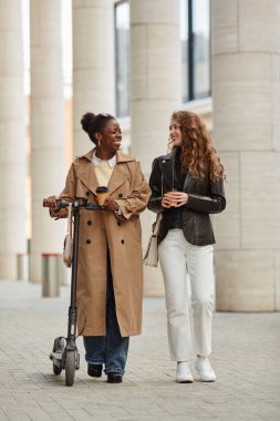 Şehirde kameraya doğru yürüyen elektrikli scooterlı iki genç kadının dikey portresi.