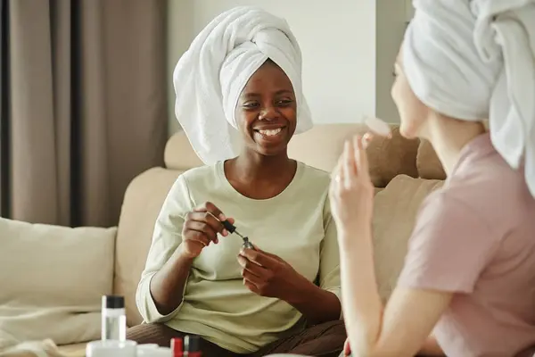 Portrait of smiling black woman with hair in towel chatting with girlfriend while enjoying self care day at home