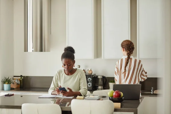 Minimal portrait of young black woman using smartphone while working from home in kitchen interior copy space
