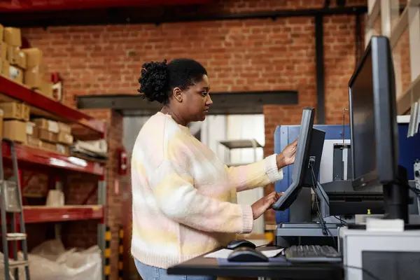 Side view portrait of adult black woman using computer while setting up printing machines in industrial shop