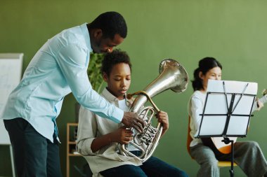 Genç kendinden emin Afrikalı Amerikalı müzik öğretmeni trompetli çalışkan öğrenciye gitar çalan genç kıza karşı