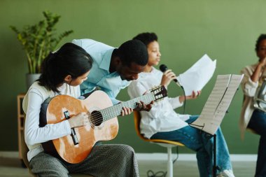 Genç Afrikalı Amerikalı erkek müzik öğretmeni genç Asyalı kız öğrencinin akustik gitarıyla eğilip ona danışıyor.