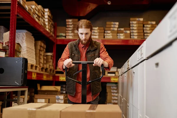 Front view portrait of bearded male worker using pallet jack while moving boxes at printing factory warehouse