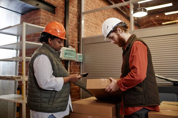 Side view portrait of two workers wearing hardhats counting boxes in factory warehouse and using tablet