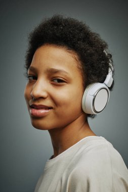 Güzel, genç ve güzel bir kız kulaklıkla müzik dinliyor ve gri arka planda tek başına dikilirken kameraya bakıyor.