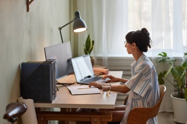 Side view portrait of young woman using laptop while working or studying at cozy home office workplace with dim lights clipart