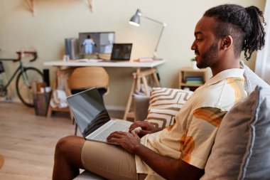 Koltukta oturup boş ekrana gülümserken dizüstü bilgisayar kullanan yaratıcı siyahi adamın yan görünüm portresi