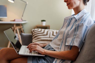 Evdeki kanepede oturup çalışırken dizüstü bilgisayar kullanan gülümseyen genç bir kadının kesilmiş portresi.