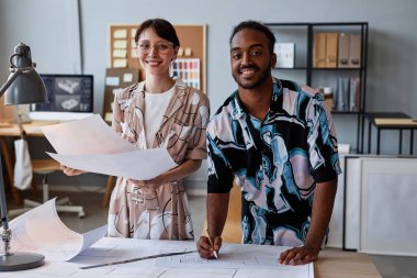 İç tasarım projesi için yer planları üzerinde çalışan ve kameraya bakan gülümseyen iki genç mimarın belden yukarı portresi