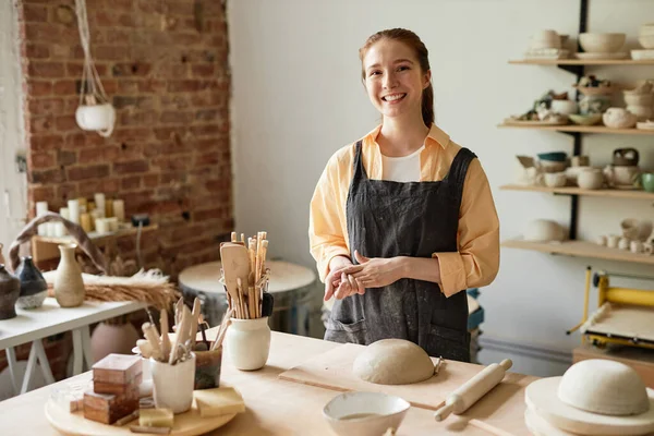Waist up portrait of smiling young woman wearing apron in cozy pottery studio, copy space