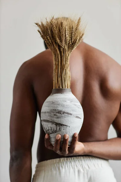 Minimal closeup of shirtless black man holding handmade ceramic vase with wheat stocks behind back, nature and wabi sabi design