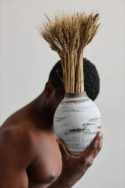 Minimal concept portrait of shirtless black man holding handmade ceramic vase with wheat stocks and hiding face, wabi sabi design