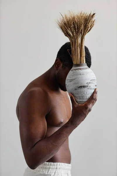 Minimal side view shot of shirtless black man holding textured ceramic vase with wheat stocks, wabi sabi design harmony with nature