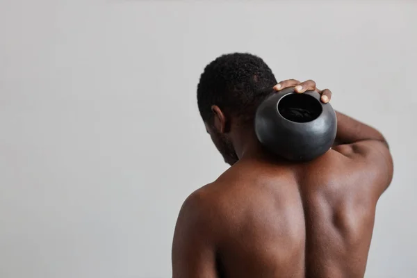 Minimal shot of shirtless black man holding round tan vase against back muscles, nature forms in design, copy space