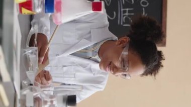 Beyaz laboratuvar önlüklü ve güvenlik gözlüklü Afrikalı Amerikalı öğrencinin dikey beliyle sınıf arkadaşlarıyla sohbet ederken okulda kimya dersi sırasında laboratuvar deneyleri yaparken...