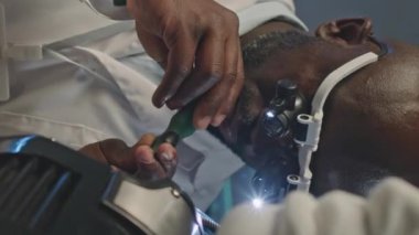 Beyaz laboratuvar önlüklü Afrikalı Amerikalı mühendisin dikey beline ve gece laboratuvarda çalışan insansı robotu tamir etmek için tornavida kullanan ışıklı büyüteçlere.