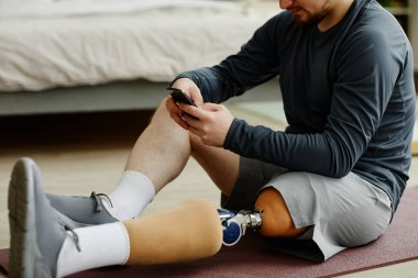 Evdeki yoga minderinde oturan ve akıllı telefon kullanan protez bacaklı adama yakın çekim.