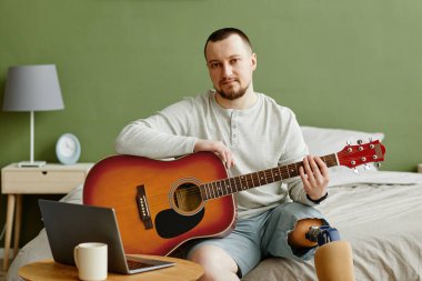 Evde gitar çalıp dizüstü bilgisayar kullanan protez bacaklı bir adamın portresi.
