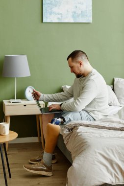 Evde yatarken dizüstü bilgisayar kullanan protez bacaklı bir adamın yan görünüm portresi.