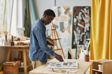 Boya fırçası ve renk paleti olan genç yaratıcı siyahi adam atölyedeki akrilik boyalarla boyanmış yeni sanat eserleri üzerinde çalışıyor.