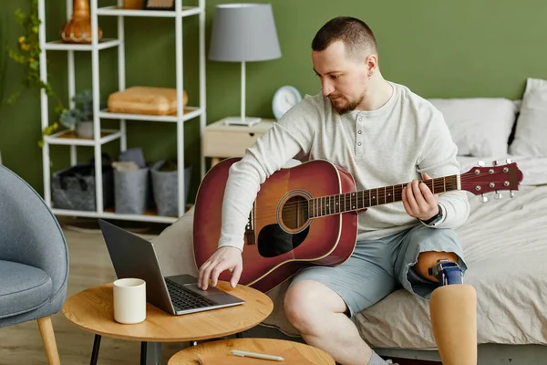Side view portrait of man with prosthetic leg playing guitar at home and using laptop for online music class, copy space