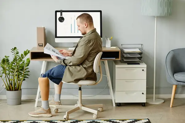 Minimal full length portrait of man with prosthetic leg working at home office and reading documents, copy space