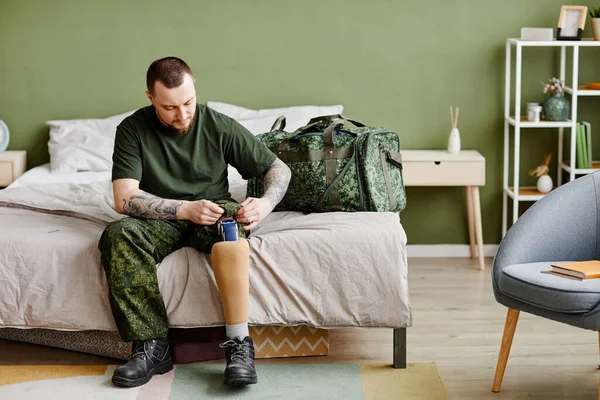 Full length portrait of military veteran with prosthetic leg putting on army uniform in bedroom, copy space
