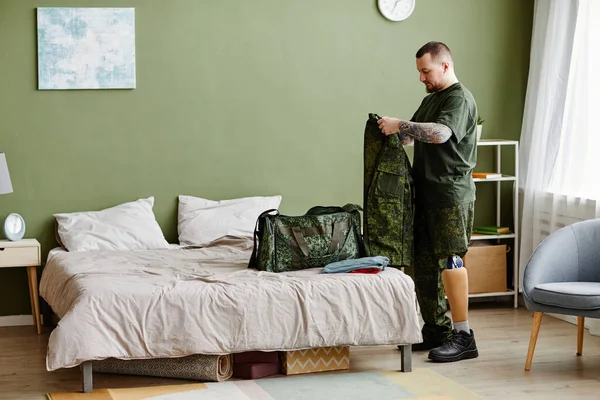 Full length portrait of military veteran with prosthetic leg packing army uniform at home, copy space