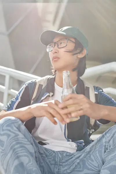 日光で屋外でストリートスタイルの服を着てガラス瓶を持っている若いアジア人男性の垂直の肖像画 — ストック写真