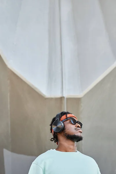 Minimal portrait of young black man wearing headphones standing by concrete wall outdoors and looking up, copy space
