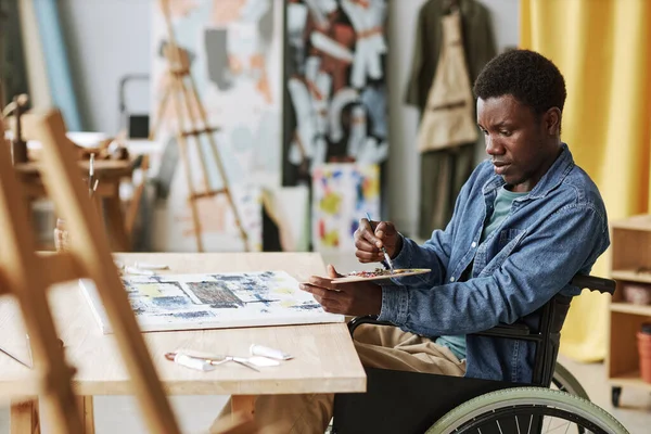 Young African American craftsman in wheelchair mixing colors on palette while sitting by table in studio of arts and painting on canvas