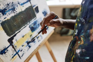 Tamamlanmamış sanat eserleriyle tuvalin önünde dururken elinde boya fırçası tutan genç Afrikalı Amerikalı ressamın yakın çekimi