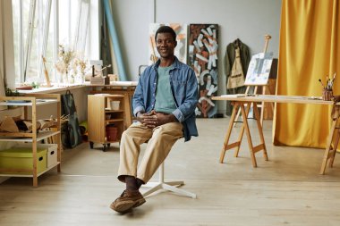 Genç profesyonel ressam ya da sanat öğretmeni geniş bir stüdyoda, sehpası, masası ve tuvalleriyle karşısında oturuyor.