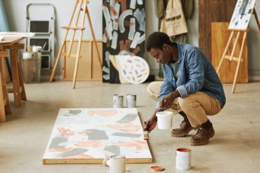 Genç siyahi bir adam çömelmiş tuvalin üzerine oturmuş ve tamamlanmamış bir sanat eseriyle onu stüdyo ya da sınıfta boyuyor.