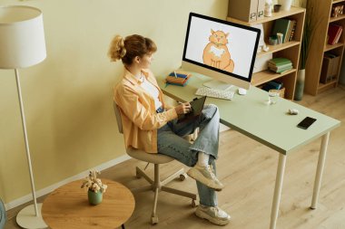 Grafik tabletli ve kalemli genç sarışın bir kadının sandalyede oturup kızıl kedi çizerken bilgisayar ekranına bakması.