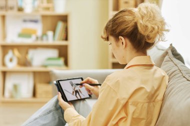 Evdeki kanepede otururken tablet ekranda savaşçının grafiksel resmini çizen aleti ve stili olan genç sarışın kadının arka görünüşü.