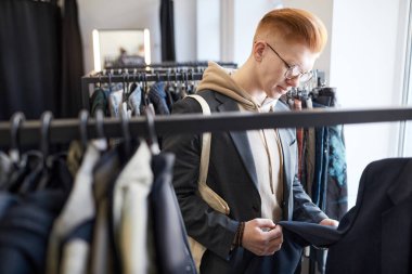 Genç kızıl saçlı adamın ikinci el dükkanından alışveriş yaparken askılarda kıyafetlerini karıştırması.