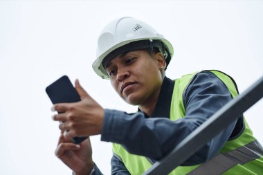 Düşük açılı kadın işçinin portresi. Kasket takıyor ve akıllı telefonu gökyüzüne karşı kullanıyor.