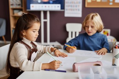 Modern anaokulu ya da anaokulu sınıfında masa başında oturmuş pastel boyayla kağıt çizen iki çalışkan öğrenci.